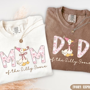 May include: Two Comfort Colors t-shirts, one ivory and one espresso, with the text "MOM of the Silly Goose" and "DAD of the Silly Goose". Each shirt features a goose graphic wearing a party hat and a necklace.