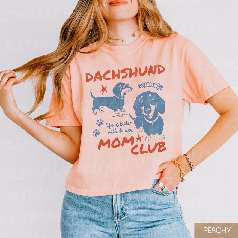 May include: Peach-colored t-shirt featuring a dachshund graphic with the words "DACHSHUND MOM CLUB" in red. The shirt also includes the phrase "life is better with doxies" and illustrations of two dachshunds.