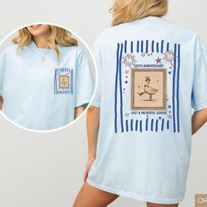 May include: Pale blue t-shirt featuring a patriotic goose design. The back displays a framed illustration of a goose in a top hat and holding a cane, with text reading "250th Anniversary" and "Just a Patriotic Goose."