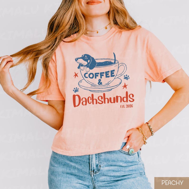 Peut inclure: T-shirt p&ecirc;che avec un motif graphique repr&eacute;sentant un teckel dans une tasse &agrave; caf&eacute;. Le texte indique "Coffee & Dachshunds est. 2026" en bleu et rouge. Le motif comprend des empreintes de pattes et des &eacute;toiles.