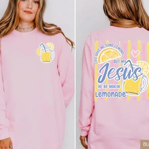 May include: A light pink long-sleeve shirt with a lemonade graphic on the front and back. The front features a yellow lemonade jar with lemons. The back has the text "Life gave me some lemons but my Jesus he be makin' lemonade."