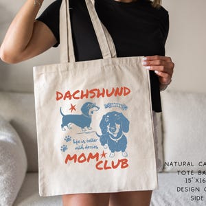 May include: Natural canvas tote bag with a dachshund design. The bag features blue illustrations of dachshunds, paw prints, and the text "DACHSHUND MOM CLUB". The tote bag measures 15" x 16".