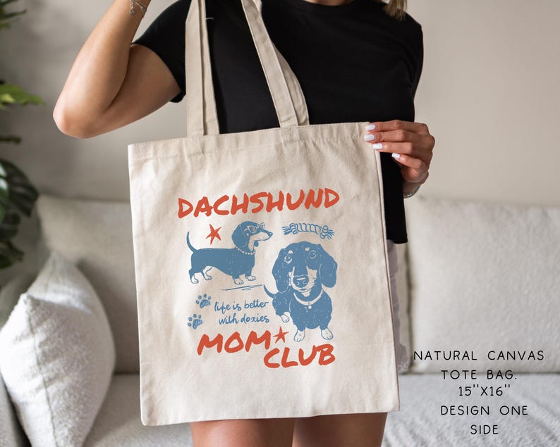 May include: Natural canvas tote bag with a dachshund design. The bag features blue illustrations of dachshunds, paw prints, and the text "DACHSHUND MOM CLUB". The tote bag measures 15" x 16".
