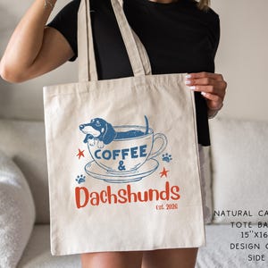 Peut inclure: Un sac fourre-tout en toile de couleur naturelle avec un motif bleu et rouge. Le motif repr&eacute;sente un teckel dans une tasse &agrave; caf&eacute;, avec les mots "Coffee & Dachshunds" et "Est. 2026". Le sac mesure 38 cm x 41 cm.