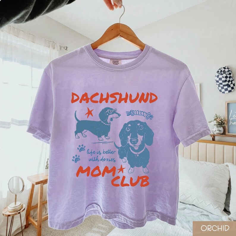 May include: A light purple t-shirt with a dachshund graphic and the words "DACHSHUND MOM CLUB". The graphic features two dachshund illustrations, paw prints, and the phrase "life is better with doxies".