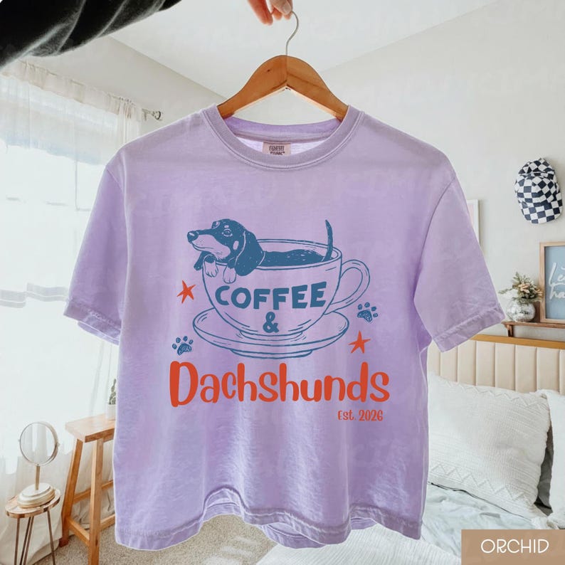 Peut inclure: Un t-shirt lilas avec un motif de teckel dans une tasse &agrave; caf&eacute;. Le texte "Coffee & Dachshunds" est imprim&eacute; en orange et bleu, avec "est. 2026" en dessous. Le t-shirt est suspendu &agrave; un cintre en bois.