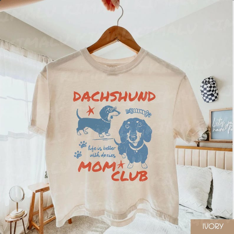 May include: Ivory-colored t-shirt with a dachshund graphic. The design features two blue dachshund illustrations, the word "DACHSHUND" in red, and the text "life is better with doxies MOM CLUB". The shirt is hanging on a wooden hanger.