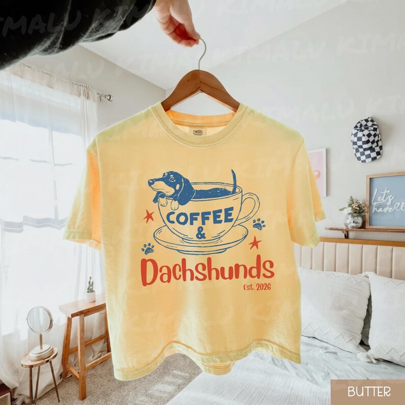 Peut inclure: T-shirt jaune avec un motif d'un teckel bleu dans une tasse &agrave; caf&eacute;. Le texte "Coffee & Dachshunds est. 2026" est en rouge et bleu. Le t-shirt est suspendu &agrave; un cintre en bois.