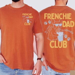 May include: Burnt orange t-shirt with "FRENCHIE DAD CLUB" printed on the front and back. The back graphic features a French Bulldog wearing sunglasses, drinking from a cup, and the phrase "life is better with frenchies."