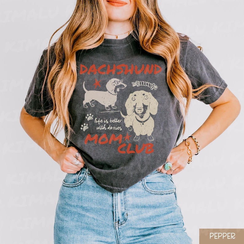 May include: Dark gray t-shirt featuring a dachshund graphic with the words "DACHSHUND MOM CLUB" in red. The shirt also includes the phrase "life is better with doxies". The design showcases two dachshund illustrations.
