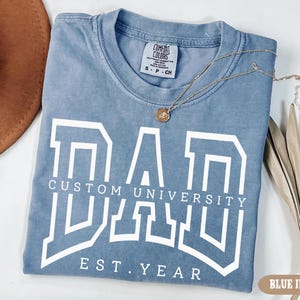 May include: A light blue t-shirt with a white graphic that says "DAD CUSTOM UNIVERSITY EST. YEAR" in a collegiate font.