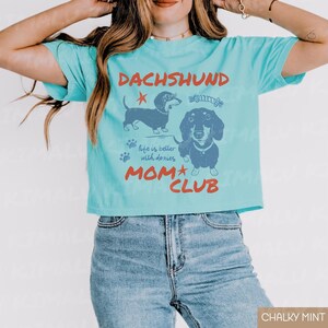 May include: A light blue t-shirt with a graphic featuring two dachshund illustrations and the text "DACHSHUND MOM CLUB." The shirt also includes the phrase "life is better with doxies."