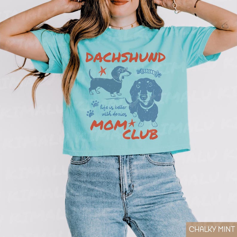 May include: A light blue t-shirt with a graphic featuring two dachshund illustrations and the text "DACHSHUND MOM CLUB." The shirt also includes the phrase "life is better with doxies."