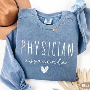 May include: A blue jean-colored sweatshirt with the words "PHYSICIAN associate" in white script, along with a small heart graphic. A gold necklace and a brown hat are also visible.