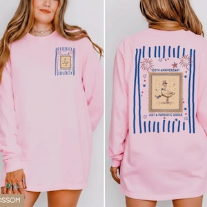 May include: A pale pink long-sleeved top featuring a goose in a hat graphic, with the text "Just a Patriotic Goose" and "250th Anniversary". The design is framed by blue stripes and stars.