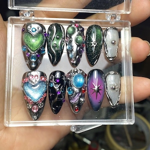 May include: A set of ten press-on nails in a clear plastic case, featuring almond-shaped tips with metallic silver accents, colorful 3D gems, heart-shaped charms, starburst motifs, and gradient finishes in shades of green, blue, purple, and black.