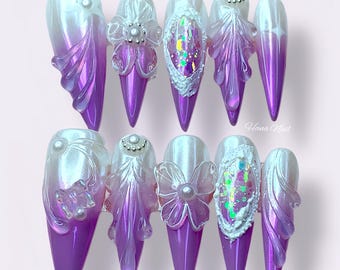 May include: A set of ten long, pointed artificial nails. Each nail features a gradient of white to purple, with intricate 3D embellishments including flowers, pearls, and iridescent accents. The nails are arranged on a light pink surface.