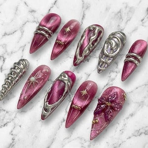 May include: A set of ten press-on nails featuring a deep magenta cat-eye finish. The nails are adorned with various silver metallic 3D accents, including swirling liquid-like shapes, star charms, small gold beads, and a sculpted floral design.