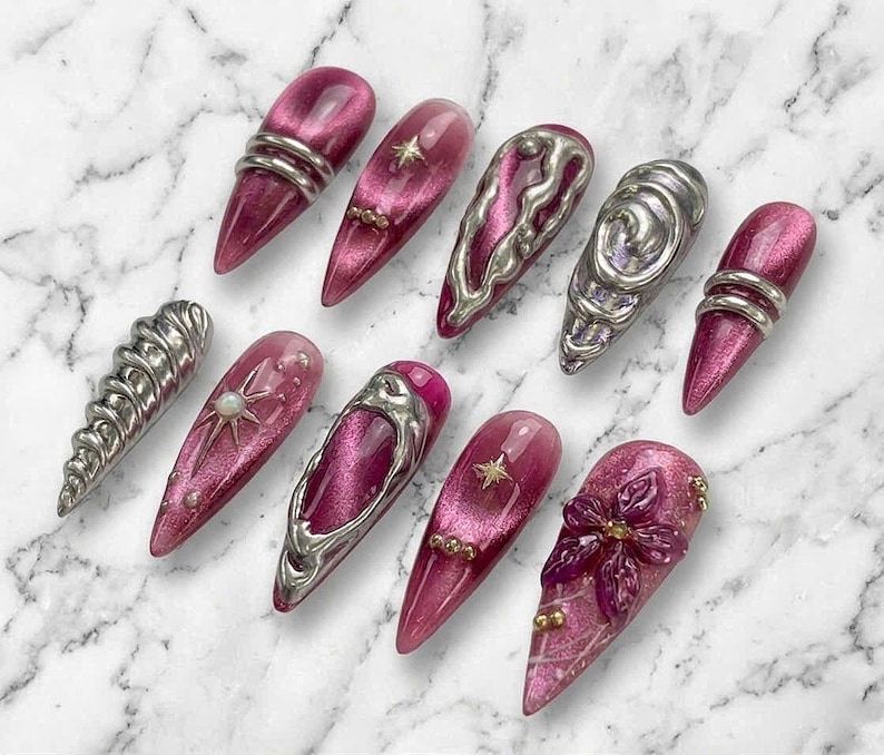May include: A set of ten press-on nails featuring a deep magenta cat-eye finish. The nails are adorned with various silver metallic 3D accents, including swirling liquid-like shapes, star charms, small gold beads, and a sculpted floral design.