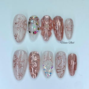 May include: A set of ten artificial nails in various designs. The nails feature a mix of clear, pink, and rose gold colors. Some nails have snowflake, bow, and star designs, while others have textured or gem embellishments. The text "Hana Nail" is visible.