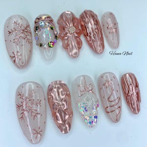 May include: A set of ten artificial nails with a winter holiday theme. The nails feature a variety of designs, including snowflakes, a Christmas tree, and a decorative ornament. The color palette includes pink, clear, and rose gold.