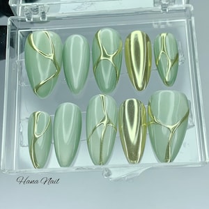 Pode incluir: Um conjunto de dez unhas postiças em forma de amêndoa numa caixa de plástico transparente. As unhas são de cor verde pálido com desenhos abstratos metálicos dourados. O texto "Hana Nail" está impresso na parte inferior esquerda da caixa.