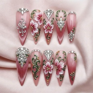 May include: Ten stiletto-shaped press-on nails featuring a pink gradient base. The designs include hand-painted white and pink flowers, green swirling vines, gold metallic accents, and clusters of clear rhinestones arranged in various patterns.