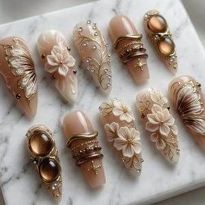 May include: A collection of ten artificial nails with various designs. The nails feature a nude base color with intricate embellishments, including gold filigree, rhinestones, and 3D floral accents. Some nails have a butterfly wing design.