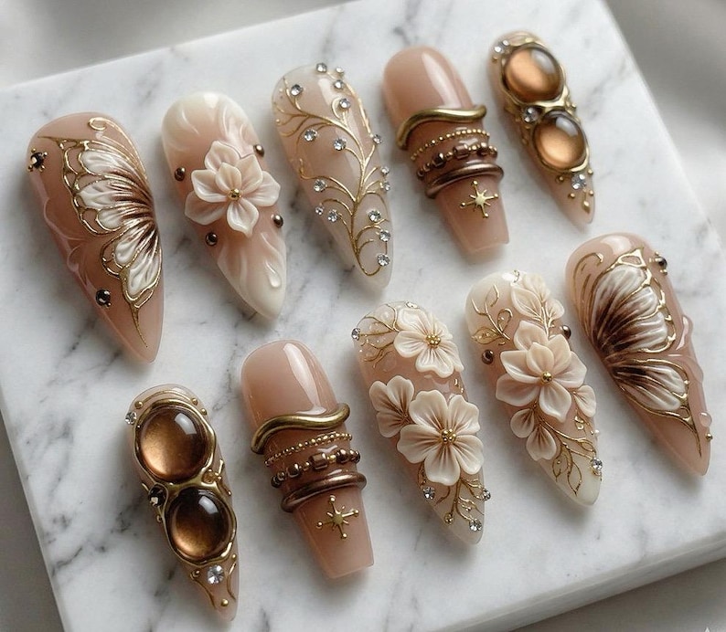 May include: A collection of ten artificial nails with various designs. The nails feature a nude base color with intricate embellishments, including gold filigree, rhinestones, and 3D floral accents. Some nails have a butterfly wing design.