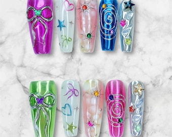 Puede incluir: Un conjunto de diez uñas artificiales en varios colores y diseños. Las uñas presentan adornos como lazos, estrellas, corazones y espirales. Los colores incluyen rosa, verde, azul y plateado, con un acabado brillante. Cada uña tiene un diseño diferente.