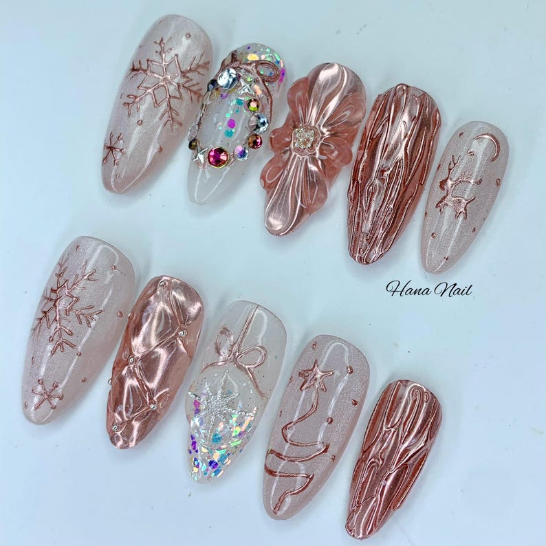 May include: A set of ten artificial nails with a winter holiday theme. The nails feature a variety of designs, including snowflakes, bows, and textured patterns in shades of pink, clear, and rose gold. Some nails are adorned with rhinestones. The text "Hana Nail" is visible.