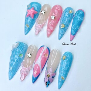 Puede incluir: Un conjunto de diez u&ntilde;as postizas largas y puntiagudas con varios dise&ntilde;os. Algunas u&ntilde;as est&aacute;n pintadas con una base azul claro y tienen un dise&ntilde;o de remolino blanco, mientras que otras est&aacute;n pintadas con una base rosa claro y tienen un dise&ntilde;o de remolino blanco. Algunas u&ntilde;as tienen un dise&ntilde;o 3D, como una estrella de mar, una flor o una concha. Las u&ntilde;as est&aacute;n dispuestas en dos filas sobre un fondo blanco. El texto "Hana Nail" es visible en la imagen.