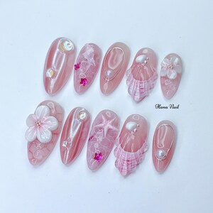 Peut inclure: Art de l'ongle rose et blanc avec un design de coquillage, d'&eacute;toile de mer et de fleur. Les ongles sont longs et de forme ovale. Le texte "Hana Nail" est visible sur l'image.