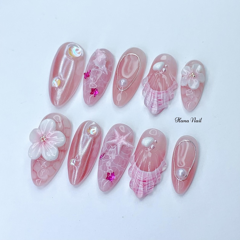 Peut inclure: Art de l'ongle rose et blanc avec un design de coquillage, d'&eacute;toile de mer et de fleur. Les ongles sont longs et de forme ovale. Le texte "Hana Nail" est visible sur l'image.