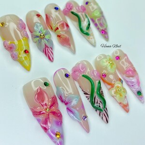 May include: A set of ten long, almond-shaped artificial nails. Each nail features a nude base with intricate floral designs in pink, yellow, and green, accented with small, colorful gems. The text "Hana Nail" is visible.