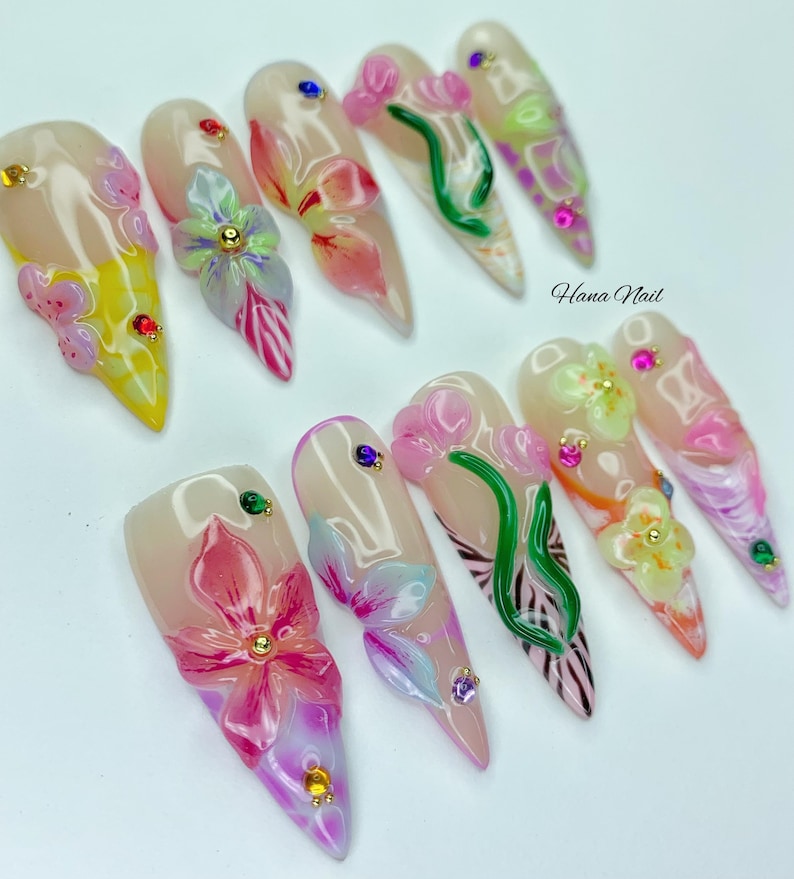May include: A set of ten long, almond-shaped artificial nails. Each nail features a nude base with intricate floral designs in pink, yellow, and green, accented with small, colorful gems. The text "Hana Nail" is visible.