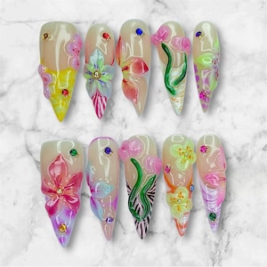 May include: A set of ten long, almond-shaped artificial nails. Each nail features a unique 3D floral design in shades of pink, yellow, and green, accented with small gemstones. The nails have a glossy finish and a natural base color.