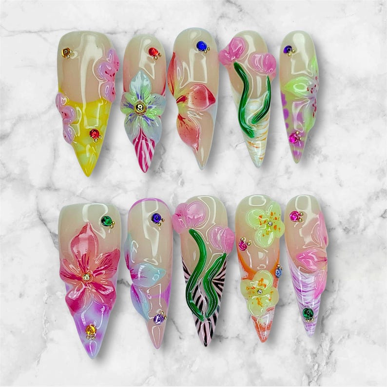 May include: A set of ten long, almond-shaped artificial nails. Each nail features a unique 3D floral design in shades of pink, yellow, and green, accented with small gemstones. The nails have a glossy finish and a natural base color.