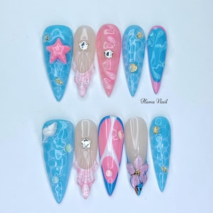 Puede incluir: Diez u&ntilde;as postizas largas y puntiagudas, azules y rosas, con varios dise&ntilde;os. Los dise&ntilde;os incluyen una flor rosa, una estrella de mar azul, un patr&oacute;n de cuadros blancos y rosas y un patr&oacute;n de remolino azul y blanco. Las u&ntilde;as est&aacute;n decoradas con diamantes de imitaci&oacute;n y purpurina dorada.