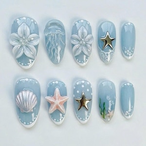 May include: A set of ten light blue press-on nails with various ocean-themed designs. The designs include 3D flowers, a jellyfish, a seashell, a starfish, gold stars, and aquatic plants. Each nail has a white border with small dots.
