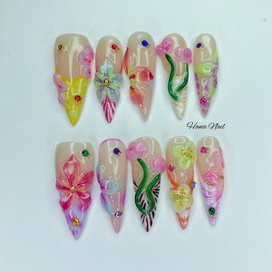 May include: A set of ten long, almond-shaped artificial nails. Each nail features a nude base with 3D floral designs in various colors, including pink, yellow, and green. Some nails have rhinestones. The text "Hana Nail" is visible.