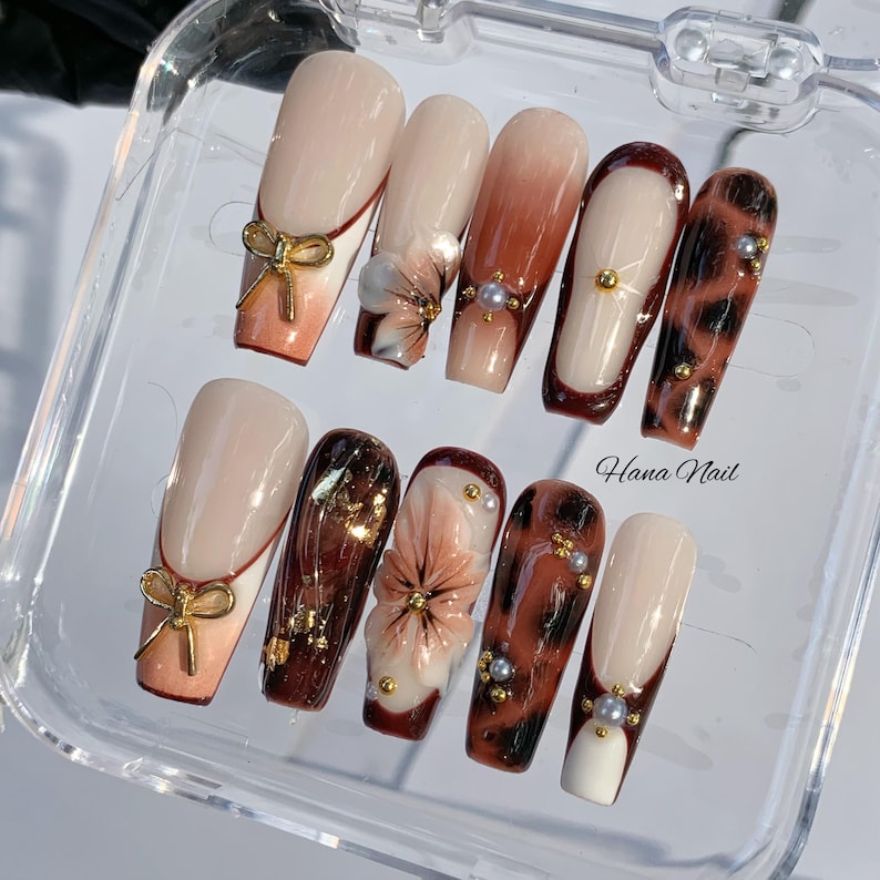 May include: A set of ten artificial nails with various designs. Some nails feature a nude base with gold bow accents, while others showcase floral and leopard print patterns. The nails are displayed in a clear plastic container. The text "Hana Nail" is visible.
