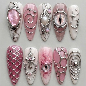 May include: A set of ten almond-shaped press-on nails featuring pink and white bases with 3D silver metallic accents. Designs include a pink gemstone, swirling silver lines, a pearl, a cat-eye, stars, a cross with chains, and scales.