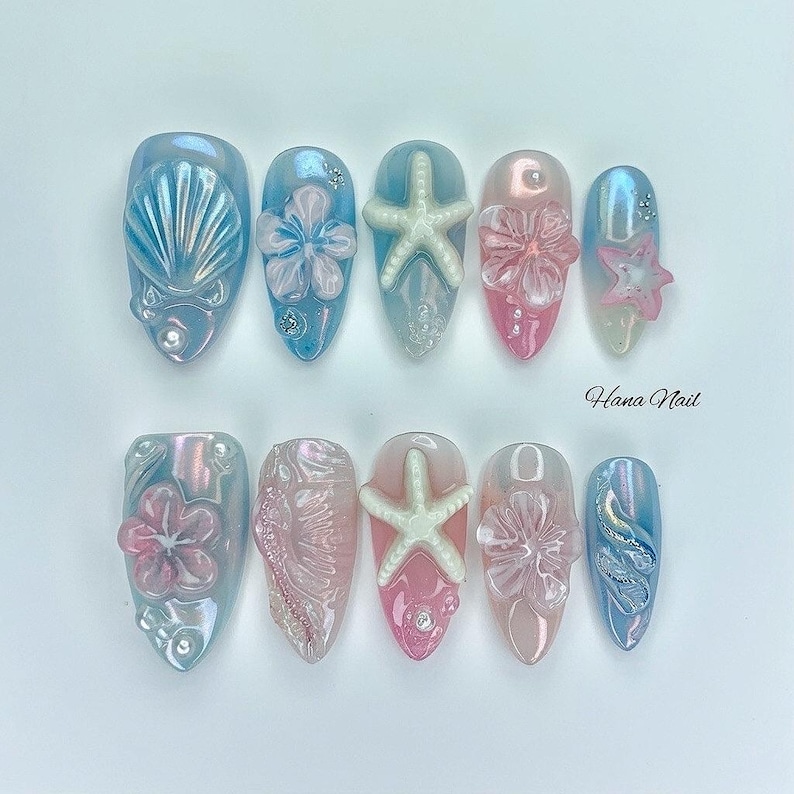 May include: A set of ten artificial nails with a summer theme. The nails feature various designs, including seashells, starfish, and flowers, in shades of blue, pink, and white. Some nails have iridescent finishes and small embellishments. The text "Hana Nail" is visible.
