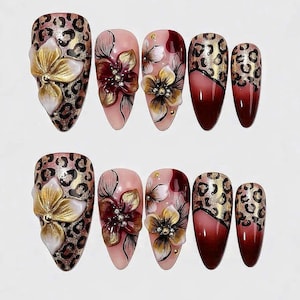 May include: A set of almond-shaped false nails. Designs include leopard print, gold and white floral accents, and burgundy ombre. Some nails feature 3D floral embellishments for a textured look. The nails are arranged in two rows.
