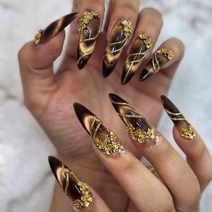 May include: Long, pointed stiletto-shaped nails featuring a dark brown cat-eye magnetic polish finish. Each nail is decorated with metallic gold wire accents that wrap around the surface and small, three-dimensional gold flower embellishments.