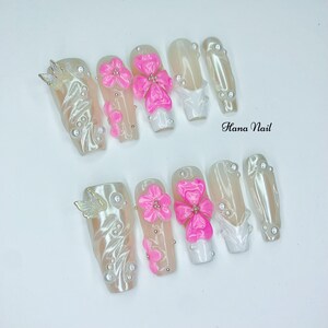 Puede incluir: Un conjunto de diez u&ntilde;as artificiales con un acabado rosa claro y nacarado. Algunas u&ntilde;as presentan detalles de flores y lazos rosas, mientras que otras tienen adornos de mariposas y perlas. El texto "Hana Nail" es visible.