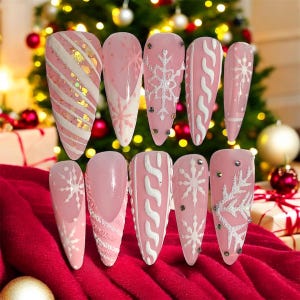 May include: A set of ten artificial nails in various designs. The nails are primarily pink with white accents, featuring snowflake patterns, candy cane stripes, and cable knit designs. Some nails have small, decorative rhinestones. The background includes a Christmas tree and red fabric.
