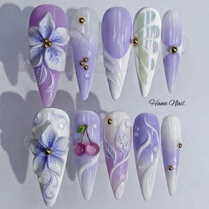 Pode incluir: Uma coleção de dez unhas artificiais longas em formato de amêndoa com designs intrincados. As unhas apresentam uma paleta de branco, lavanda e roxo, com detalhes como flores 3D, cerejas e enfeites dourados. O texto "Hana Nail" é visível.