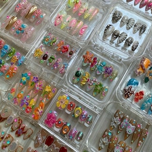 May include: Multiple clear plastic storage cases containing sets of press-on nails. The nails feature diverse designs including 3D floral patterns, starfish, seashells, rhinestones, metallic accents, and colorful painted motifs in various shapes.